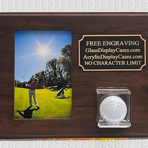 May include: A brown wooden plaque with a photo of a golfer on a green golf course. The plaque has a gold plaque with the text "FREE ENGRAVING GlassDisplayCases.com AcrylicDisplayCases.com NO CHARACTER LIMIT". A clear plastic case with a white golf ball is on the right side of the plaque.