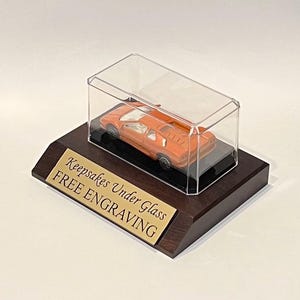 May include: An orange toy sports car enclosed in a clear rectangular display case mounted on a dark brown wooden base. A gold-colored metal plaque is attached to the front of the base with the text "Keepsakes Under Glass FREE ENGRAVING".