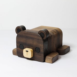 May include: A wooden bear toy lying down. The bear is made of dark brown wood with black eyes and ears. The bear's snout is a light tan colour. The toy has a simple, geometric design.