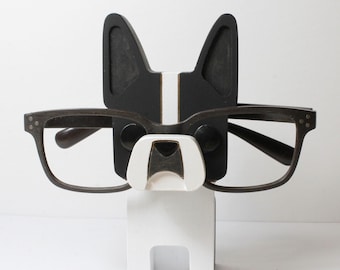 Puede incluir: Una figurita de perro de madera en blanco y negro con gafas apoyadas en su nariz. El perro tiene una raya blanca en la cara y orejas negras. La figurita es un soporte para gafas.
