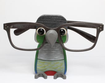Puede incluir: Un soporte de gafas con forma de loro de madera. El loro está pintado en verde, azul, gris y rojo. El loro lleva un pequeño sombrero negro y tiene un pico gris. Gafas con montura oscura descansan sobre la cabeza del loro.