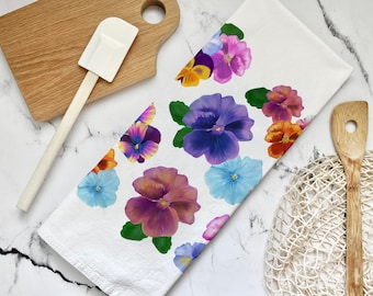 Op de afbeelding: Een witte keukendoek met een kleurrijk bloemenpatroon van paarse, blauwe, gele en oranje viooltjes. De viooltjes zijn realistisch geschilderd en hebben groene bladeren.