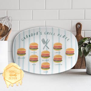 Peut inclure: Plat ovale avec un fond à rayures bleu clair et l'inscription "Grandpa's Grill Plate" en turquoise. Le plat présente des illustrations de hamburgers et des noms. Une spatule et une fourchette sont également illustrées.