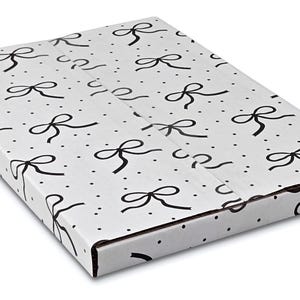 Puede incluir: Una caja rectangular blanca con un patr&oacute;n repetido de lazos negros y peque&ntilde;os puntos negros. La caja est&aacute; cerrada y parece estar hecha de cart&oacute;n, con pliegues y bordes visibles.