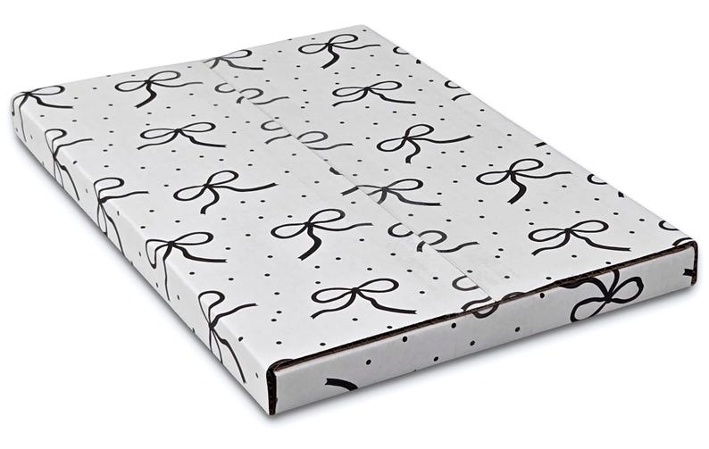 Puede incluir: Una caja rectangular blanca con un patr&oacute;n repetido de lazos negros y peque&ntilde;os puntos negros. La caja est&aacute; cerrada y parece estar hecha de cart&oacute;n, con pliegues y bordes visibles.