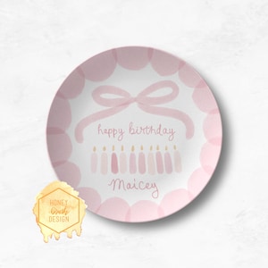 May include: A round, pink and white plate with a watercolor design. The plate features a pink bow, the words "happy birthday Maicey", and a row of candles. The plate has a scalloped pink border.