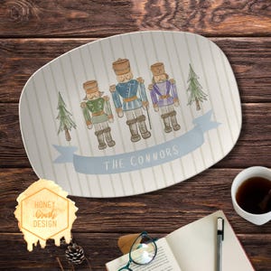 May include: Oval serving platter with a light blue and white striped background. The platter features three nutcracker figures in green, blue, and purple uniforms, with a light blue banner that reads "THE CONNORS". Two small green trees are also on the platter.