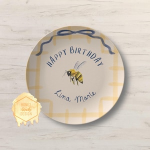 May include: A decorative plate with a yellow and white plaid pattern. The plate features a watercolor illustration of a bee and the words "Happy Birthday" and "Lina Marie" in blue script. A blue bow design decorates the top edge.