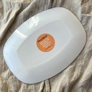 Puede incluir: Plato DecoWare rectangular blanco con esquinas redondeadas. El plato tiene un borde ligeramente elevado y un centro empotrado. Una pegatina naranja en la parte posterior dice "Microwave Safe, Dishwasher Safe, Oven Safe, Melamine Free, BPA Free."