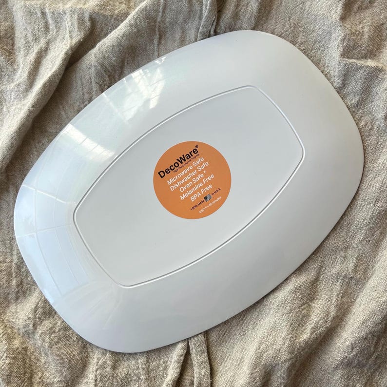 Puede incluir: Plato DecoWare rectangular blanco con esquinas redondeadas. El plato tiene un borde ligeramente elevado y un centro empotrado. Una pegatina naranja en la parte posterior dice "Microwave Safe, Dishwasher Safe, Oven Safe, Melamine Free, BPA Free."