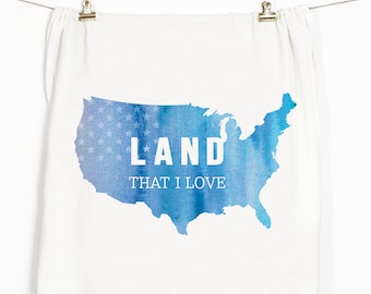 May include: A white tea towel with a blue watercolor outline of the United States. The outline is filled with white stars and the text "LAND THAT I LOVE" is printed in white.