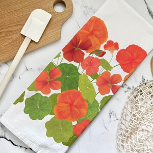 May include: A white kitchen towel with a floral design featuring orange and green nasturtium flowers. The towel is folded and displayed on a marble surface with a wooden spatula and a white mesh bag.
