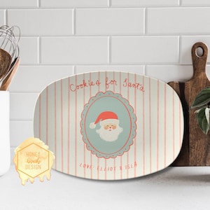 May include: An oval serving platter with pink vertical stripes and the text "Cookies for Santa." A Santa Claus graphic is in the centre, surrounded by a blue scalloped border. The text "LOVE: ELLIOT & ISLA" is below the Santa graphic.