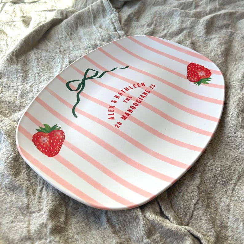 Puede incluir: Plato ovalado blanco con rayas rosas y dos fresas rojas. El plato tiene un lazo verde y el texto "ALEX & KATHLEEN THE 20 MANOOGIANS 25".