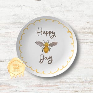 May include: White ceramic plate with a yellow bee illustration and the words "Happy Day" in brown script. The plate has a yellow wavy border. A logo with the text "Honey Brush Design" is in the lower left corner.