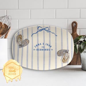 May include: Oval serving platter with a cream background and blue vertical stripes. The platter features watercolor illustrations of oysters and the text "EMMA & JOHN THE REDMONDS 20 25" in blue. A blue bow is at the top.