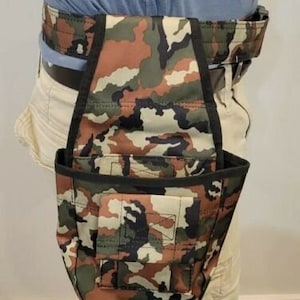 May include: A camouflage-patterned tool belt with multiple pockets and a black strap. The belt is worn around the waist and has a brown and green camouflage pattern.