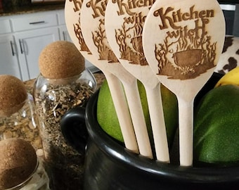 May include: Four wooden spoons with the text "Kitchen Witch" burned into the wood. The spoons are in a black cauldron with green fruit.