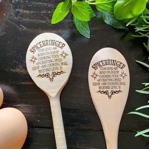 May include: Two wooden cooking spoons with the text "SPICEBRINGER" and game-like stats etched into the wood. The spoons are light-colored and are set against a dark wooden surface with fresh herbs and eggs.