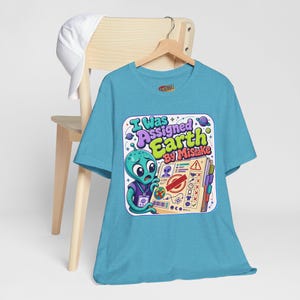 May include: A light blue t-shirt featuring a graphic of a green alien looking at a clipboard with the text "I Was Assigned Earth By Mistake". The design includes various space-themed icons, a checklist, and a red stamp. The shirt is displayed on a wooden hanger.