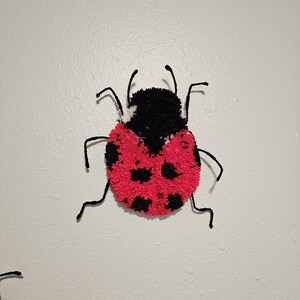 Puede incluir: Un adorno de pared de mariquita hecho a mano. La mariquita es roja con manchas negras y una cabeza negra. Tiene seis patas negras y dos antenas negras. La mariquita está hecha de hilo y está montada en una pared blanca.