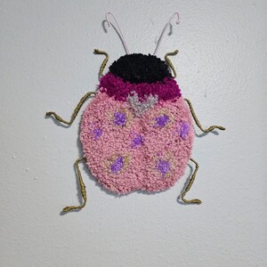 Puede incluir: Adorno de pared hecho a mano con mechones en forma de mariquita. La mariquita tiene una cabeza negra, un cuerpo rosa y morado con lunares morados y beige, y patas y antenas de alambre. El fondo es una pared blanca lisa.