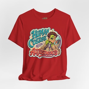 May include: A red short-sleeved t-shirt featuring a graphic illustration of a green alien pulling at its human face mask in a mirror. The text reads Human Costume Needs Adjustments in blue and red stylized lettering with small star accents.