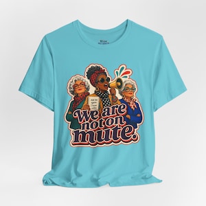 Peut inclure: Un t-shirt bleu clair avec une illustration de trois personnes, un mégaphone et un carnet. Le texte indique "Ask me again. I have notes." et "We are not on mute.". Le motif comprend des accents colorés et le logo de la marque Wise Women.