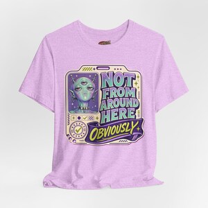 May include: A light purple t-shirt featuring a graphic of a three-eyed alien with antennae and a choker. The design includes a badge-style frame with the text "NOT FROM AROUND HERE" and a banner below with the word "OBVIOUSLY" in yellow lettering.