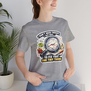 以下が含まれることがあります： 「I don't need therapy, I just need... to fix that one tiny thing」の文字が入った、グレーのTシャツ。拡大鏡、裁縫道具、糸などのグラフィックが描かれています。