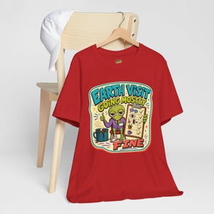 May include: A red t-shirt featuring a graphic of a green alien in a purple suit holding a clipboard with a checklist. The text reads "EARTH VISIT GOING MOSTLY FINE". The alien is sitting next to a blue mug of coffee and has a bandaged leg.