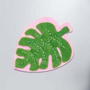 May include: A felt leaf-shaped applique in green and pink. The green leaf has a textured surface and is stitched with white thread. The leaf is outlined with a pink border, creating a layered effect. The applique is likely for crafting or decoration.