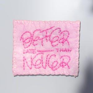 Puede incluir: Un parche rectangular rosa con la frase bordada "BETTER LATE THAN NEVER" en hilo rosa brillante. Los bordes están cosidos con hilo rosa a juego, creando un borde texturizado.