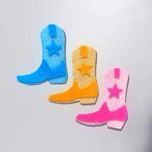 Puede incluir: Tres botas de vaquero de fieltro en azul, naranja y rosa, cada una con un adorno de estrella. Las botas están dispuestas sobre un fondo gris claro, mostrando sus colores vibrantes y su diseño lúdico. Un artículo decorativo divertido.
