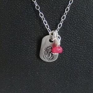 May include: A silver chain necklace with a silver pendant featuring a Ganesha design. A small, bright pink gemstone hangs from the pendant.