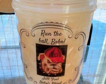 Puede incluir: Un vaso de plástico transparente y texturizado lleno de una sustancia clara. La etiqueta muestra un bulldog con una gorra roja y negra, con el texto: Run the ball, Bobo! Jett Fuel Bobo's Blend 16 oz | 473 g. Una pajita blanca sobresale.