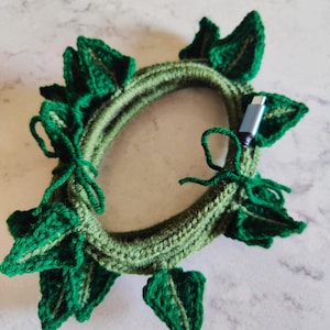 May include: A handmade, green crochet cable organizer shaped like a wreath with leaf accents. The cable organizer has a USB-C connector inserted. The leaves are a darker shade of green than the wreath.