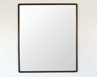 May include: Rectangular mirror with a dark brown frame. The mirror is mounted on a white wall. The frame is a thin, simple design, and the mirror's surface reflects the surrounding environment.