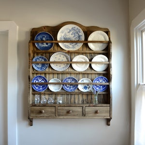 May include: A wooden plate rack with shelves and drawers, displaying a collection of decorative plates. The plates feature blue and white patterns. Small glass vases and cups are also visible.