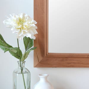 Puede incluir: Una flor blanca en un jarr&oacute;n de vidrio transparente con un jarr&oacute;n de cer&aacute;mica blanca a su lado. El jarr&oacute;n est&aacute; sobre una superficie de madera delante de un espejo con marco de madera.