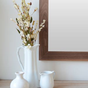 Puede incluir: Una jarra de cer&aacute;mica blanca con un ramo de flores blancas secas, junto con dos jarras de cer&aacute;mica blanca m&aacute;s peque&ntilde;as sobre una superficie de madera.