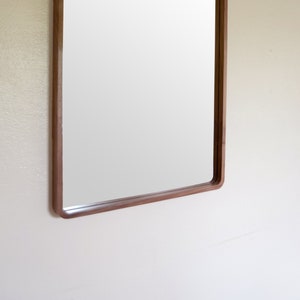 Puede incluir: Un espejo rectangular con un marco de madera marr&oacute;n. El espejo est&aacute; colgado en una pared blanca.