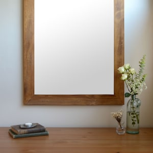 Puede incluir: Un marco de madera marrón con fondo blanco. El marco está colgado en una pared blanca. Hay flores blancas en un jarrón de vidrio y una vela pequeña sobre una mesa delante del marco.