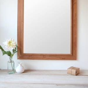 Puede incluir: Un espejo grande y rectangular con un marco de madera natural. El marco tiene un borde simple, ligeramente elevado. El espejo est&aacute; colgado en una pared blanca sobre una superficie de madera.