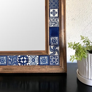 Könnte beinhalten: Rechteckiger Spiegel mit Holzrahmen. Der Rahmen ist mit blau-weiß gemusterten Fliesen verziert. Der Spiegel steht auf einer schwarzen Oberfläche, mit einem weißen Topf mit Grünpflanzen im Hintergrund.