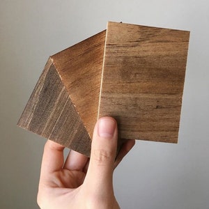 May include: A hand holds three square wooden samples, showcasing variations in wood grain and color. The samples range from light to dark brown, with visible textures. The wood appears to be unfinished, highlighting the natural patterns.