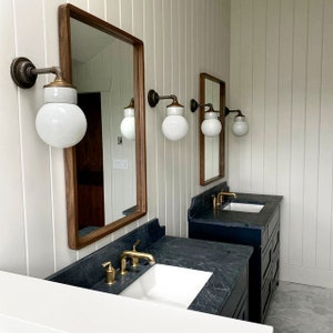 Puede incluir: Dos tocadores de ba&ntilde;o con encimeras negras y lavabos de porcelana blanca. Cada tocador tiene un grifo de oro cepillado y una l&aacute;mpara de pared con un globo de vidrio blanco.