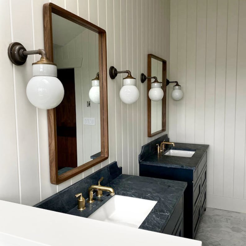 Puede incluir: Dos tocadores de ba&ntilde;o con encimeras negras y lavabos de porcelana blanca. Cada tocador tiene un grifo de oro cepillado y una l&aacute;mpara de pared con un globo de vidrio blanco.