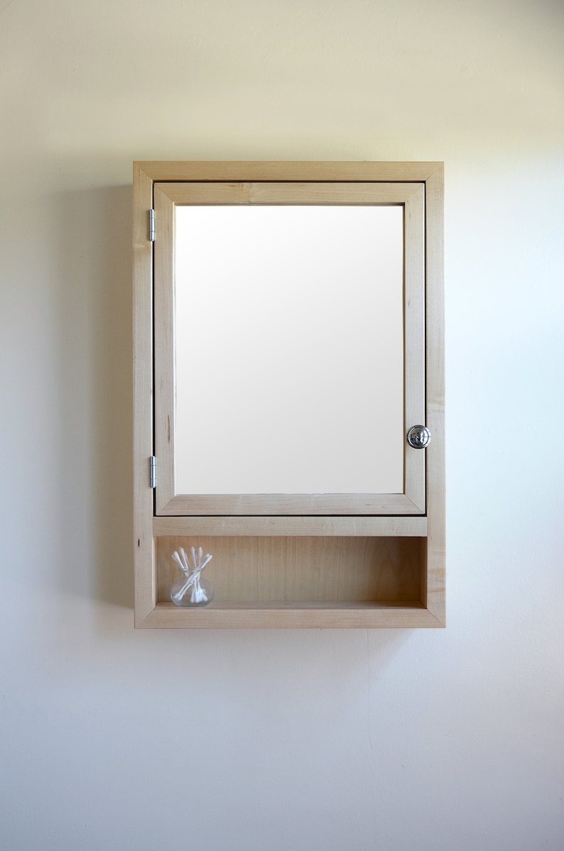 May include: A light-colored wooden medicine cabinet with a mirrored door and a small shelf. The cabinet has a silver knob and a small glass jar with cotton swabs on the shelf.