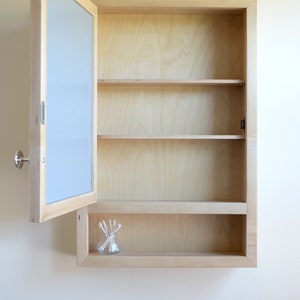 May include: A light-colored wooden medicine cabinet with a mirror door, partially open. The cabinet has three shelves and a small glass jar with cotton swabs. The cabinet is mounted on a white wall.